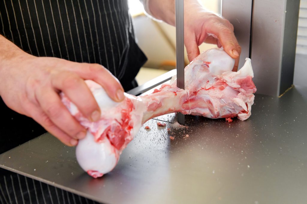 Butcher slicing a fresh trimmed raw calf femur on a band saw to make marrow bones in a close up on his hands