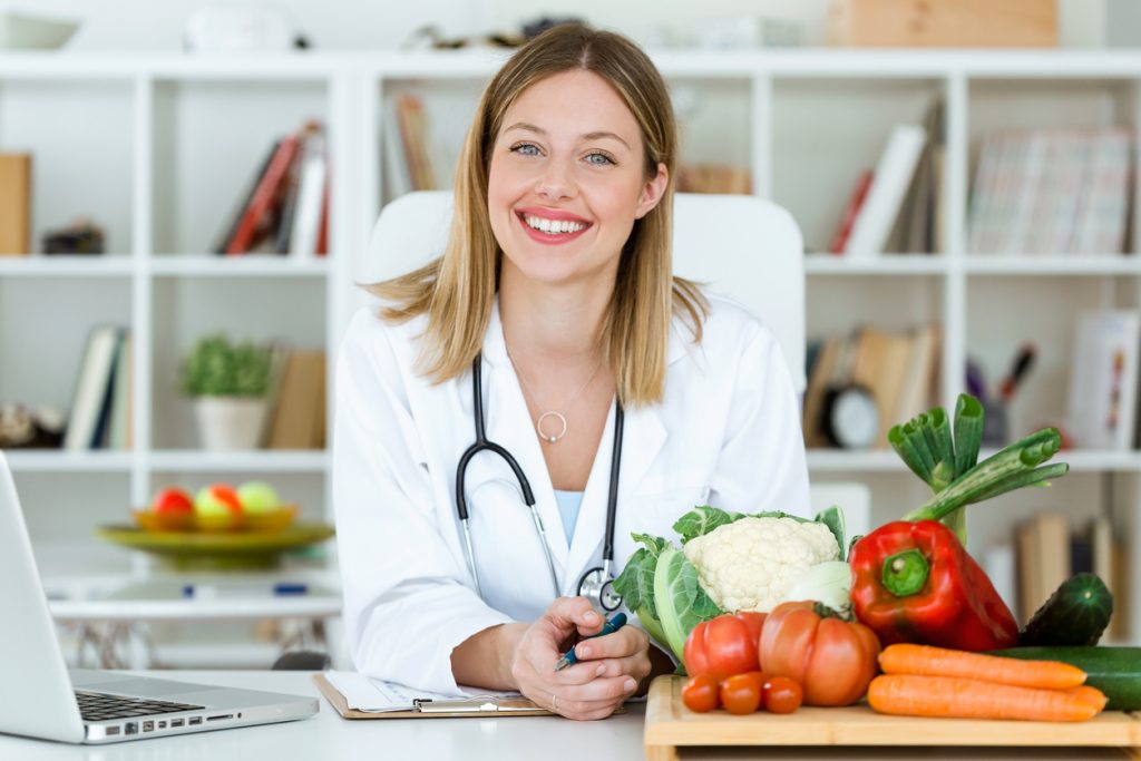 Portrait of beautiful smiling nutritionist looking at camera and showing healthy vegetables in the consultation.